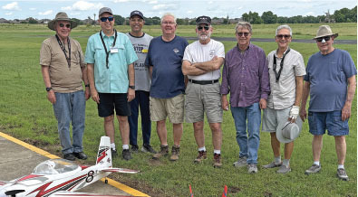Eight men stand smiling in a grassy field, model airplane on the ground.