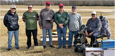 Six men standing outdoors on grass, one sitting with model airplanes and equipment.