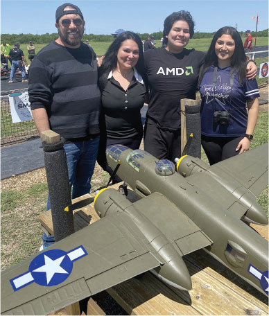 Four people smiling behind a model military plane on a wooden platform outdoors.