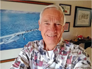 Smiling man in a patterned shirt, standing indoors with framed airplane artwork.