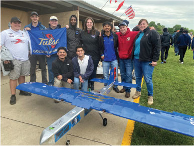Team posing with a blue model aircraft, holding a "Tulsa Golden Hurricane" banner outside.