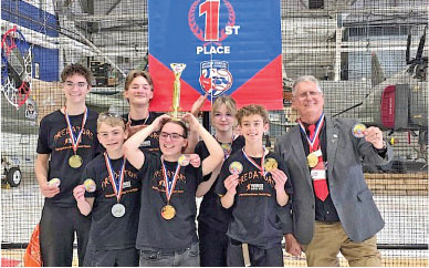 Team wearing medals celebrates first place, holding trophy. Banner in background.