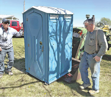 Two men standing near a blue portable toilet on grass.
