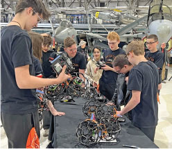 Teenagers examining robotic equipment on a table in a hangar with planes.