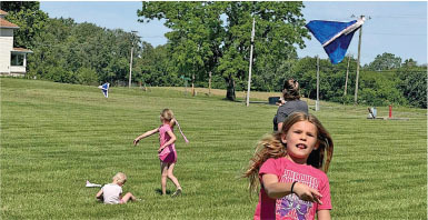 Girl flying kite on grassy field with trees and people in the background.