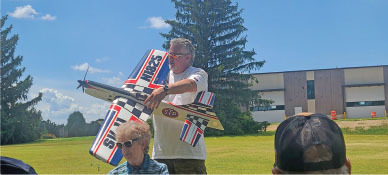 Man holding a model airplane in a grassy area with trees and a building in the background.