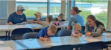 Kids and adults seated at tables coloring and writing in a bright room.