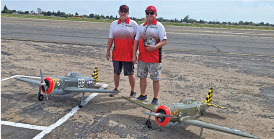 Two people in matching shirts stand on a tarmac with two large model airplanes.