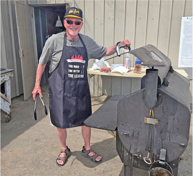 Man in apron and hat smiling, holding tongs by a large grill.