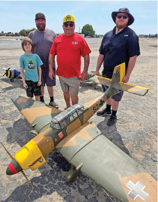 Four people stand behind a large model airplane on a sunny day.