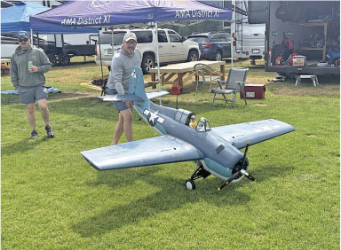 Remote-controlled plane on grass with people nearby, tents and vehicles in the background.