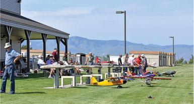 Outdoor gathering with people and model airplanes on grass, mountains in background.