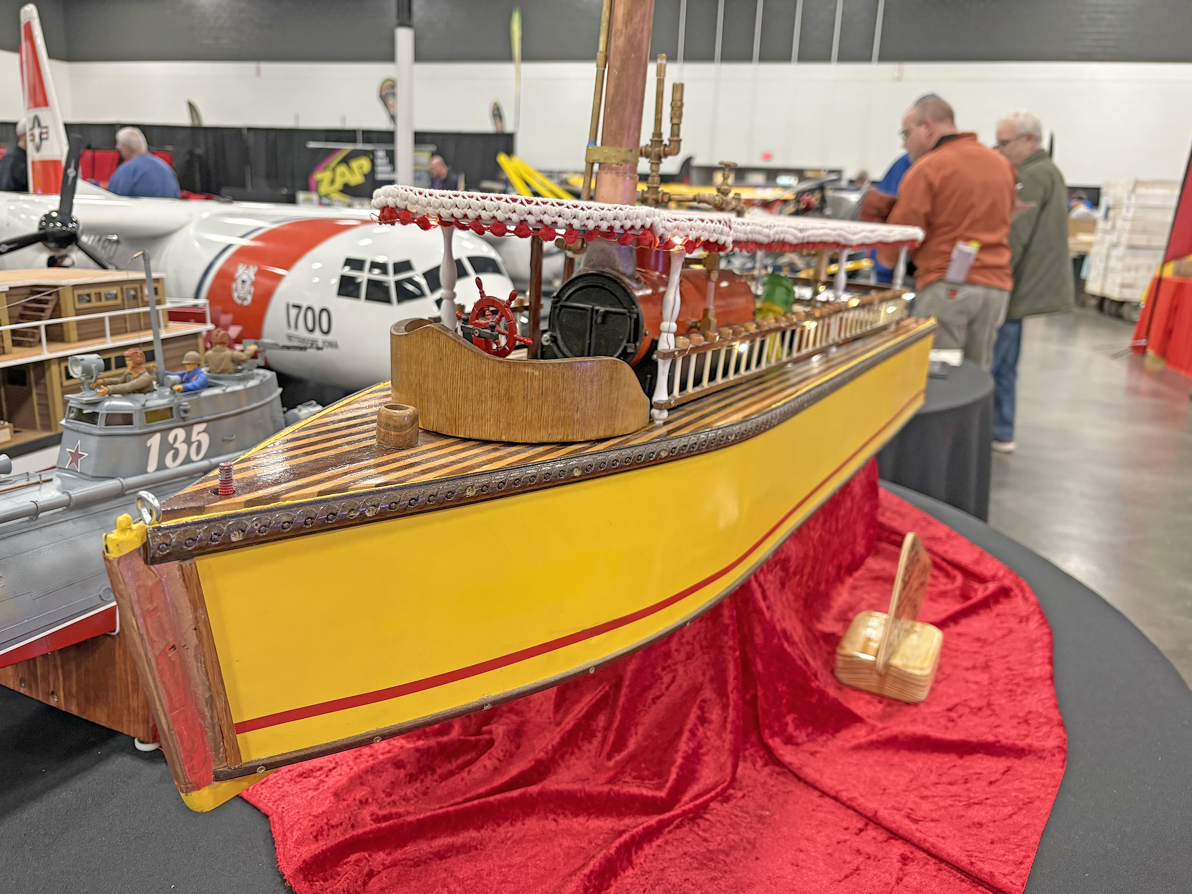 Model boat with yellow hull on red cloth, indoor exhibition setting.