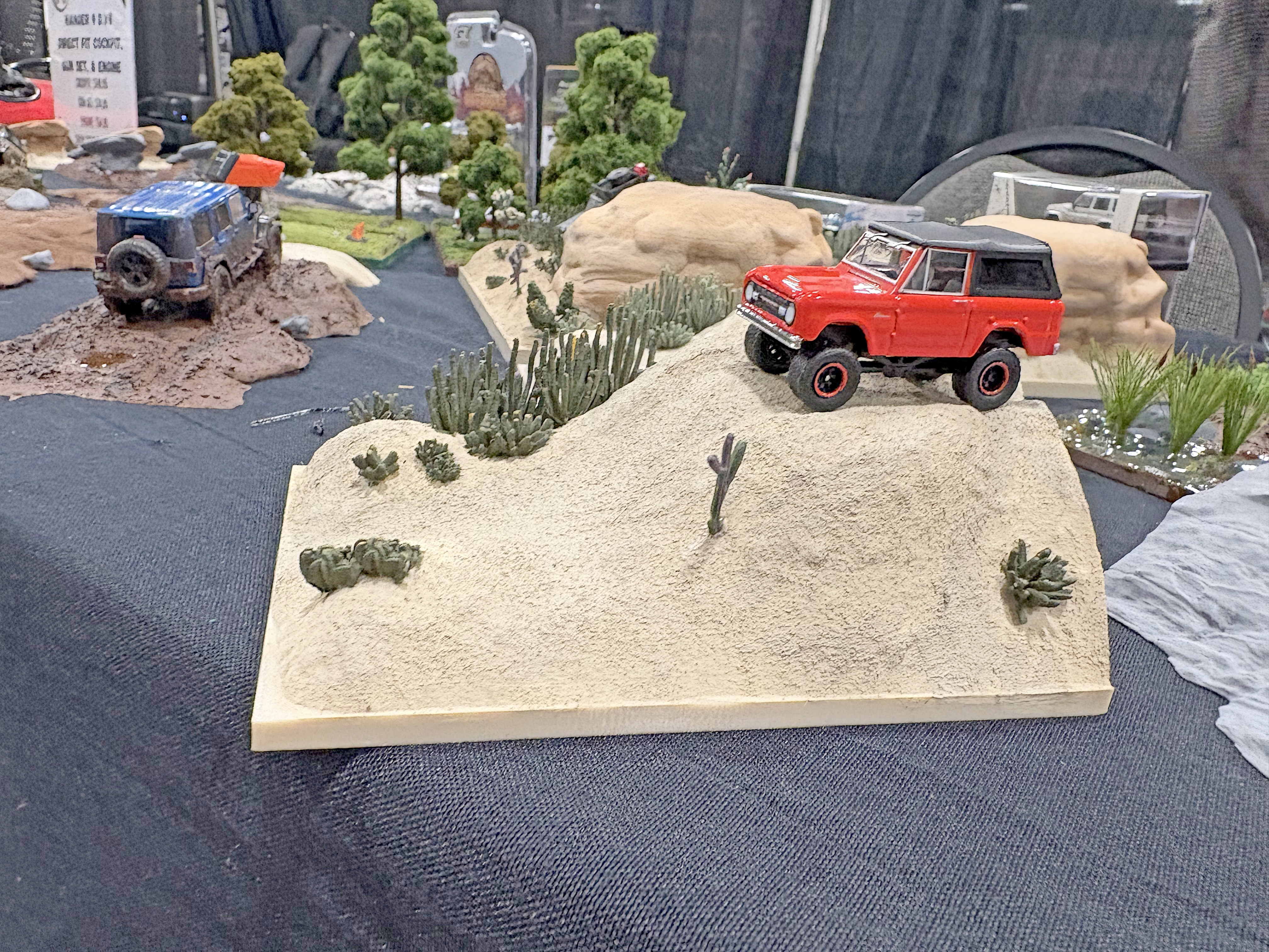 Red toy SUV on a sandy hill diorama, surrounded by small shrubs.