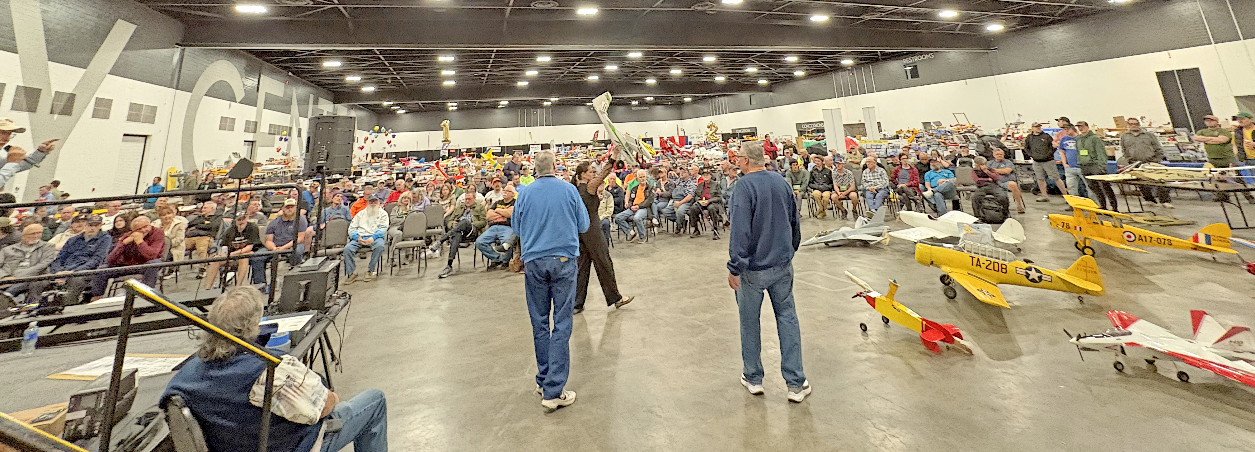 Large indoor event with people seated and model airplanes on the floor.