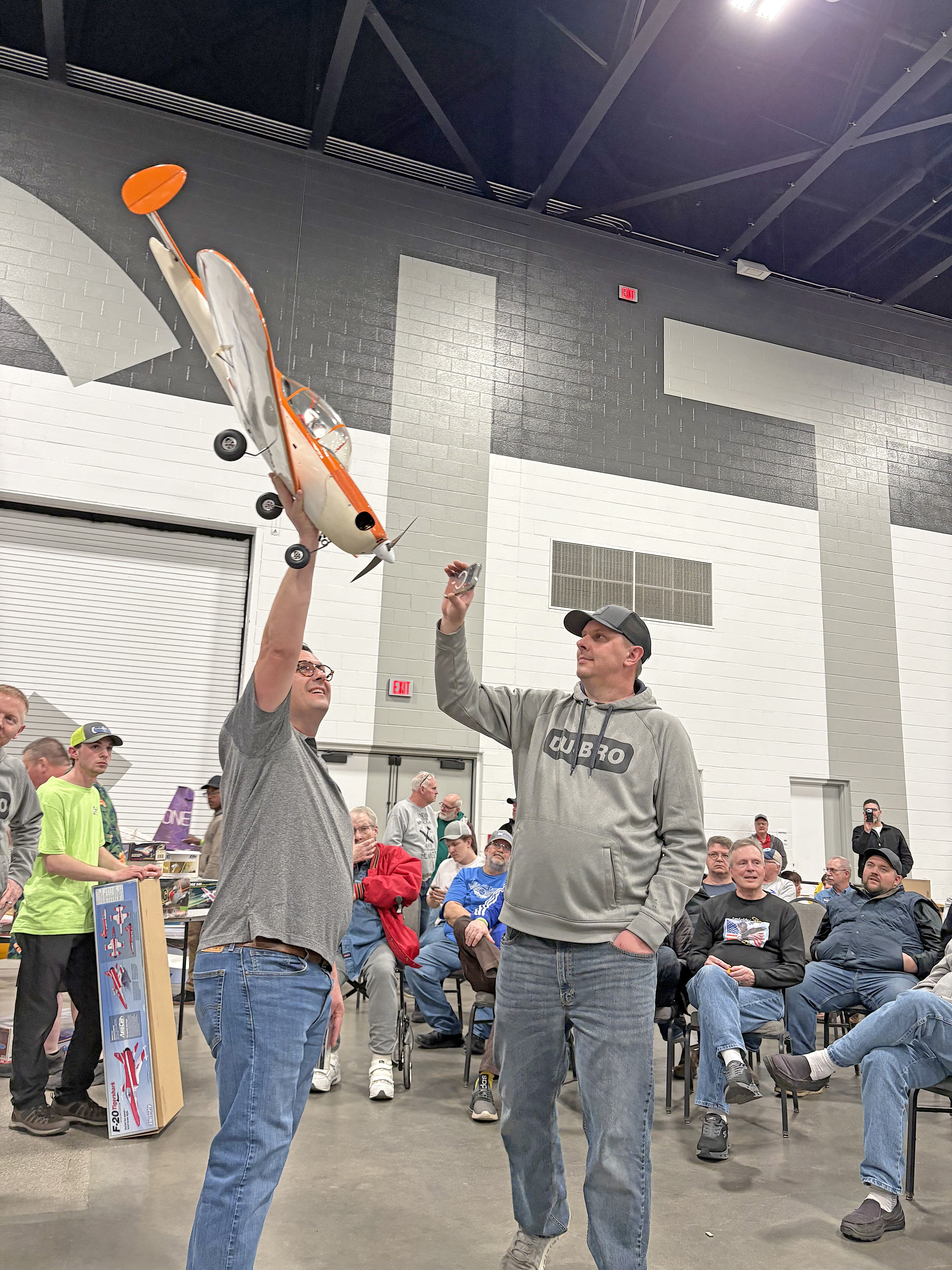 Two men in a hall holding a model plane, surrounded by seated spectators.