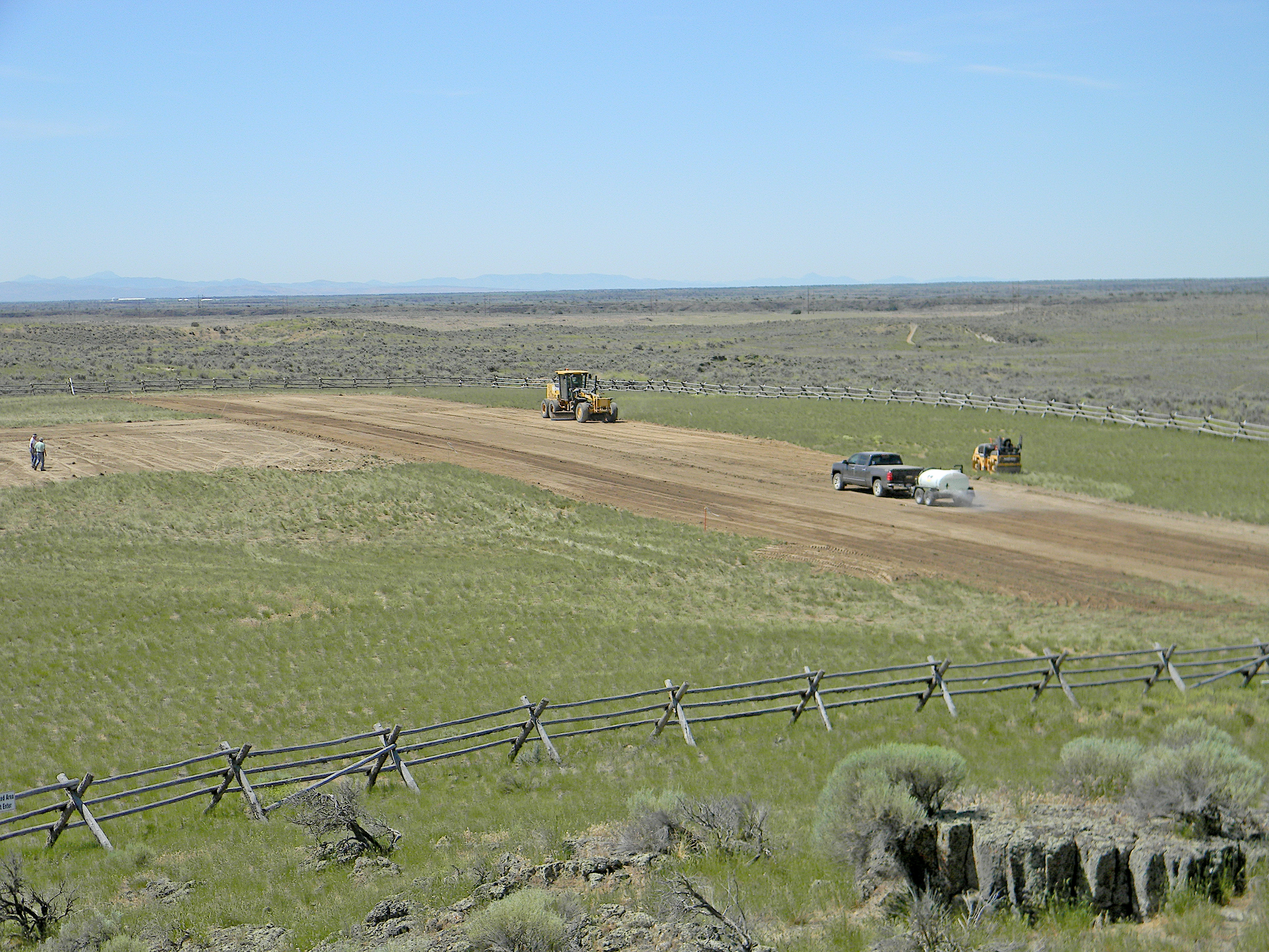 Construction vehicles on a dirt road in a grassy field, with a wooden fence in foreground.