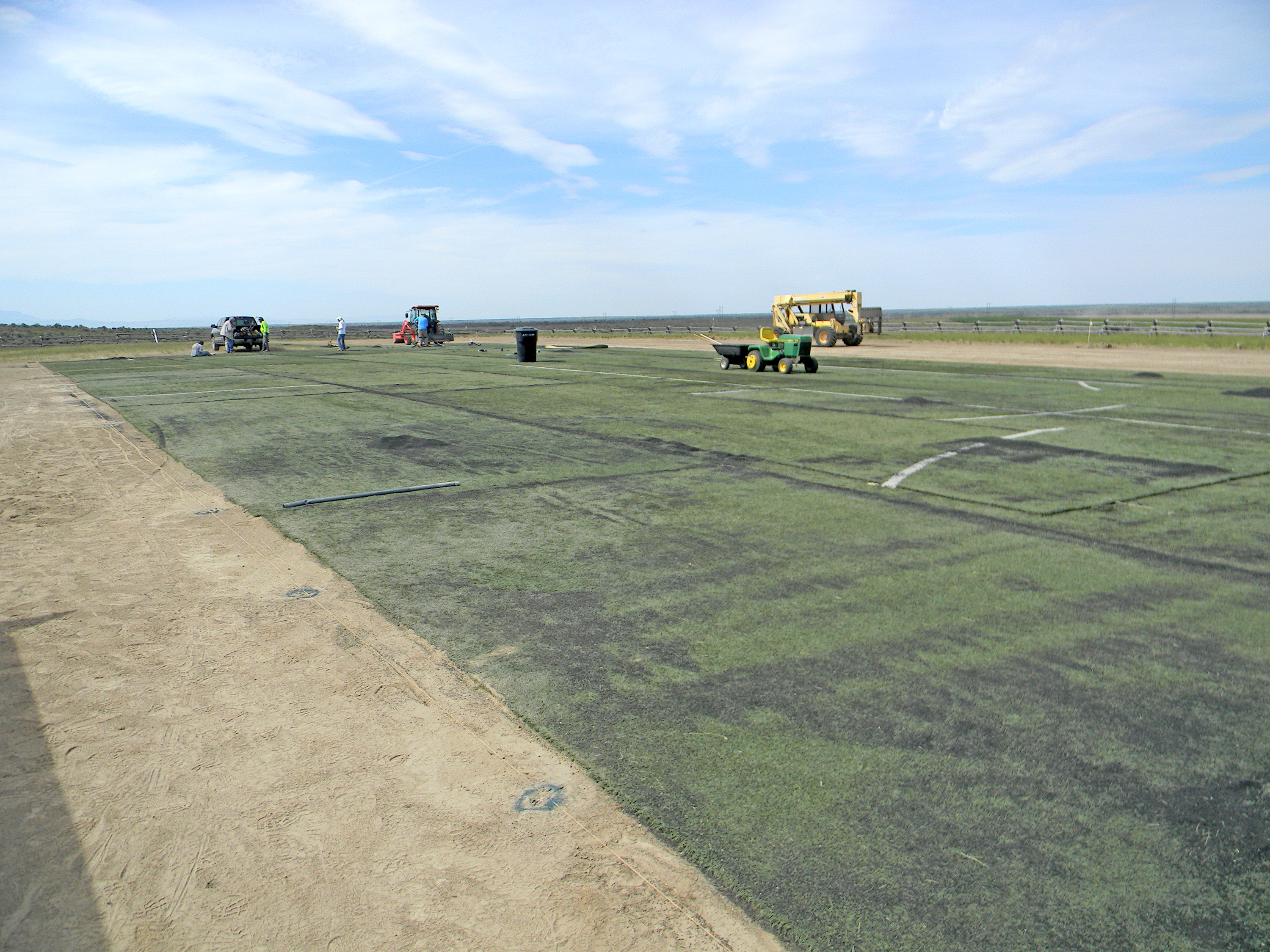 Construction vehicles on a large, green and sandy field under a blue sky.