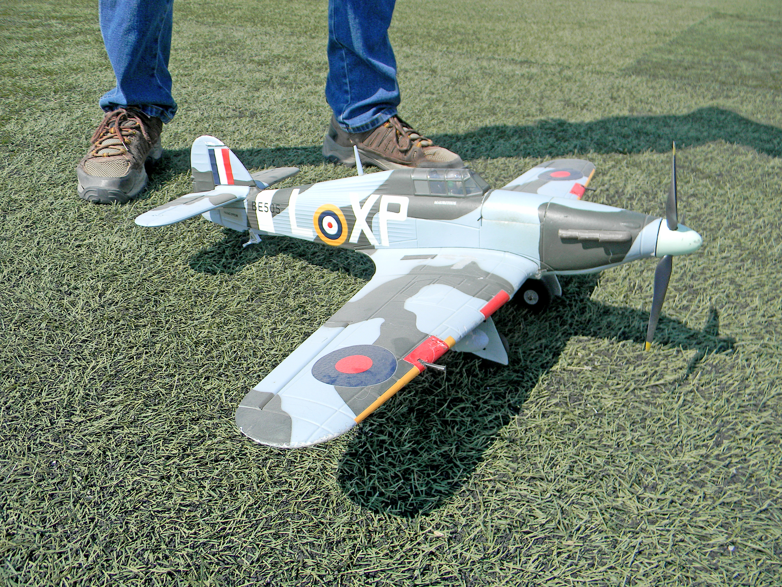Model airplane on grass with a camouflage pattern, near someone’s feet in brown shoes.