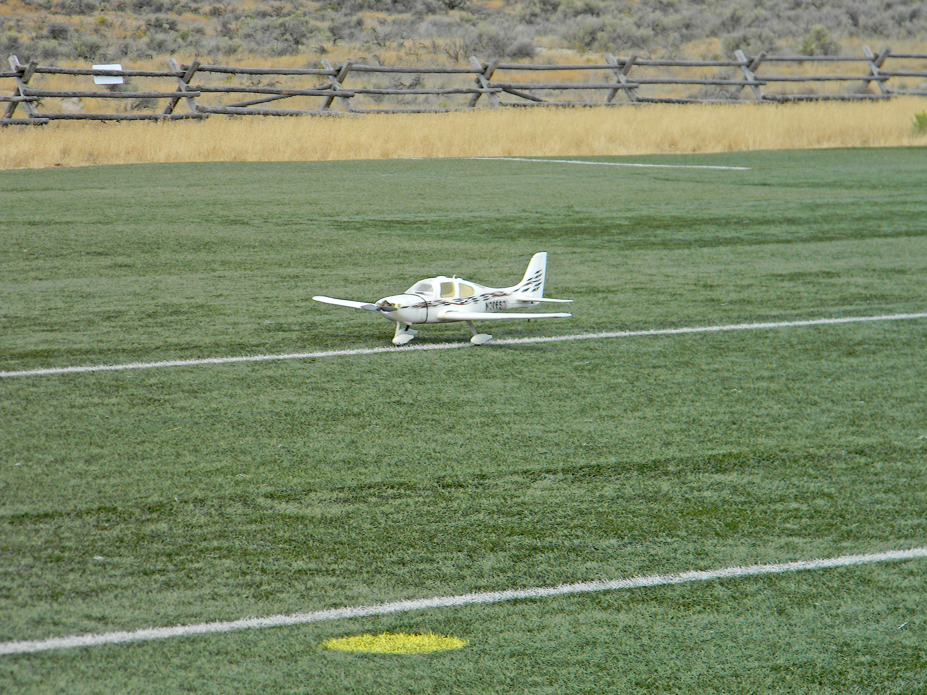 Remote-control plane on a grassy field, with a wooden fence in the background.
