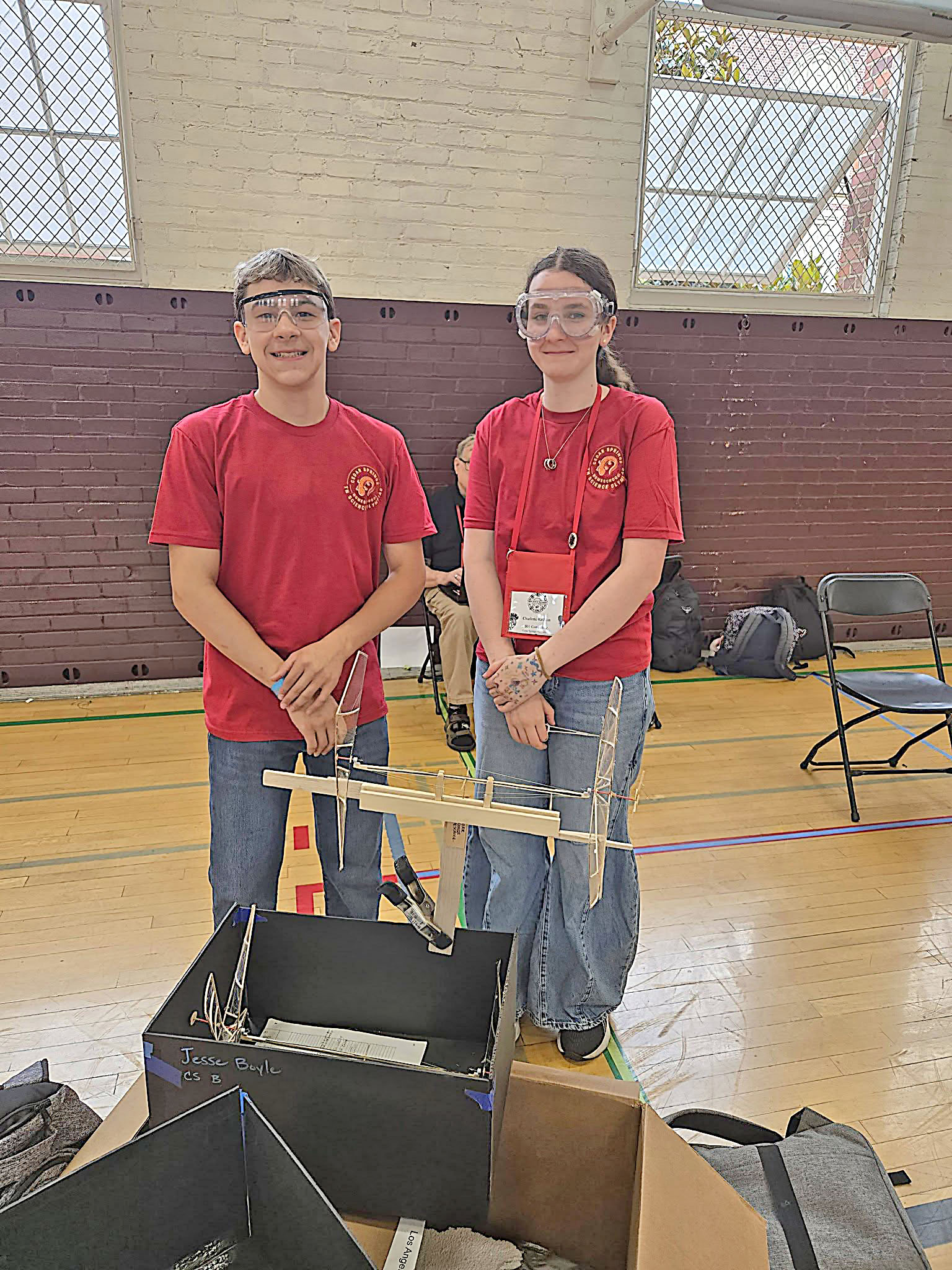 Two students in red shirts and safety goggles, standing in a gym with equipment.