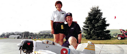 Two people smiling with a model airplane outside, next to a tree and green lawn.