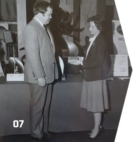 Two people exchange something, standing in front of a counter; number "07" in corner.