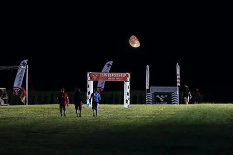 Three people walking on grass at night under a moonlit sky, near a finish line.