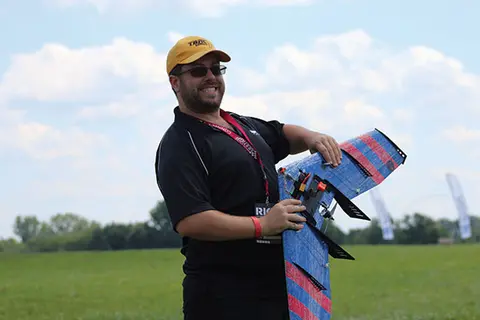 Man holding a blue and red model airplane outdoors, wearing a yellow cap.