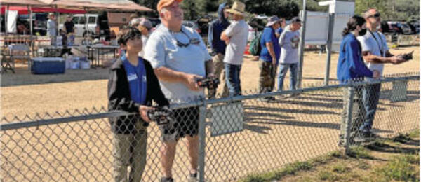 People stand behind a fence, watching an outdoor event under a sunny sky.