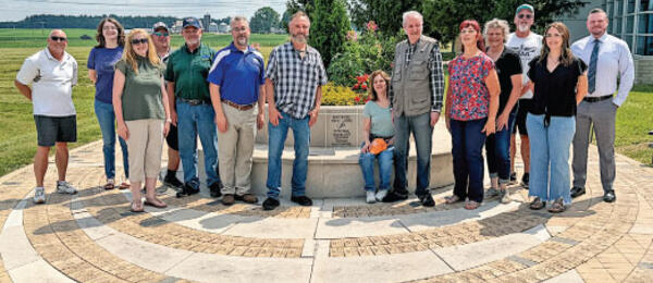Group of people standing outdoors on patterned stone path, smiling in front of a plaque.