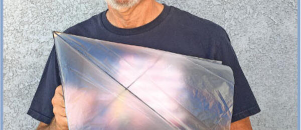 Smiling man holding a small, translucent kite in front of a textured gray wall.
