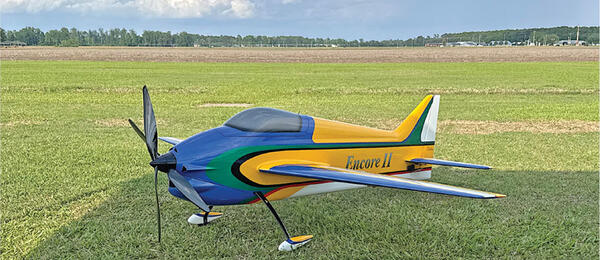 Colorful model airplane labeled "Encore II" on grass with cloudy sky.
