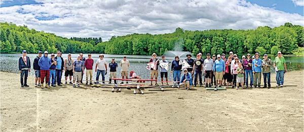 Group of people on a sandy beach, trees and a lake in the background.