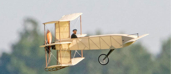 Historical biplane model in flight with blurred trees in the background.
