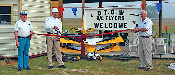 Three men hold a large ribbon in front of model planes and a welcome sign.