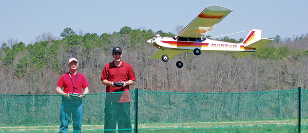 Two people fly a model airplane outdoors behind a green fence.