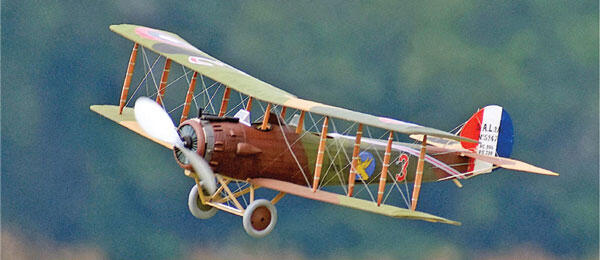 Vintage biplane with a wood frame and colorful markings in flight against a green backdrop.