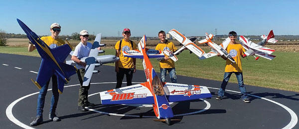 Five people hold large model aircraft on an outdoor runway, smiling at the camera.