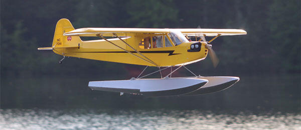 Yellow seaplane flying low over a calm lake with trees in the background.