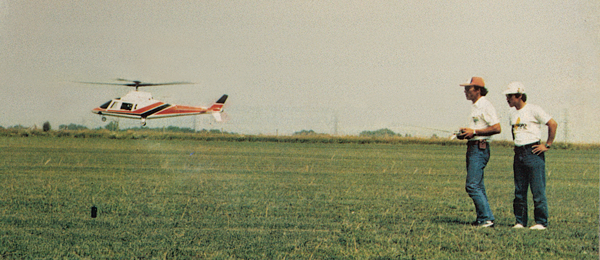 Model helicopter hovering, two people watching on a grassy field.