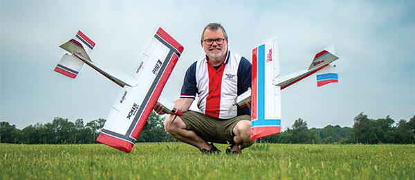 Man crouching with model airplane on grass, overcast sky.