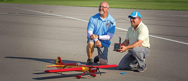 Two men kneeling beside two red and yellow model airplanes on a runway.