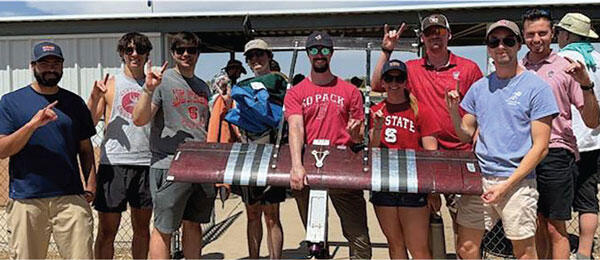 Group holding a small aircraft model outdoors, smiling and wearing casual clothes.