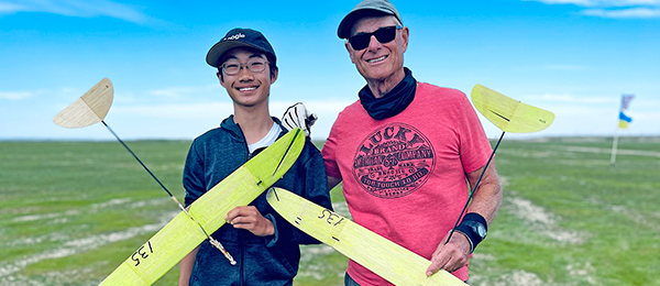 Two people holding yellow model gliders, smiling on a grassy field.