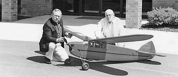 Two men kneel beside a large model airplane outdoors.
