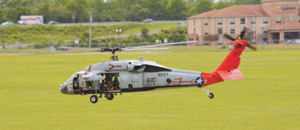 Helicopter in flight over grassy field, with distant buildings and trees.