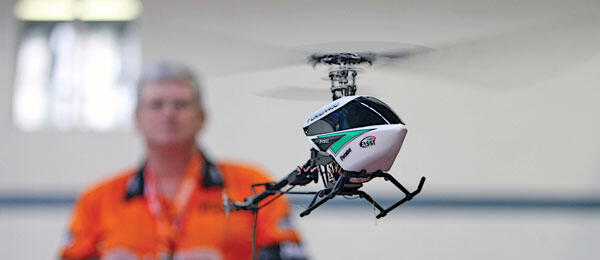 Small helicopter drone flying indoors, person in orange shirt blurred behind.