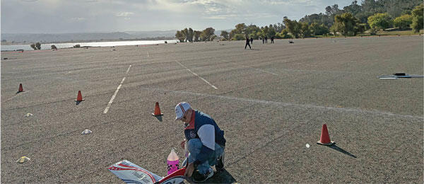 Man crouched on large asphalt area, surrounded by traffic cones, with trees in the background.