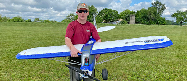 Man in sunglasses holding a large blue and white model airplane on a grassy field.